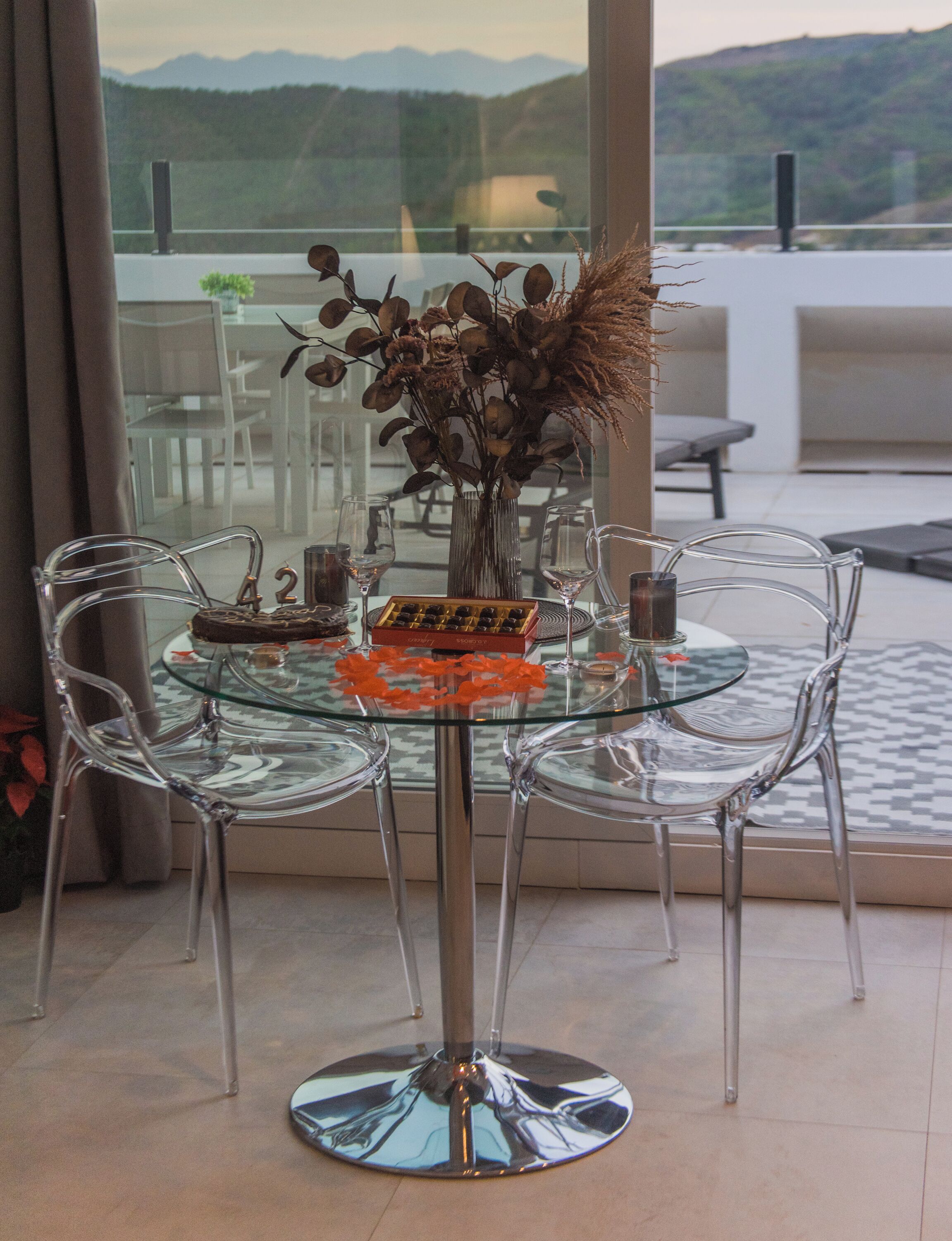 Una mesa con cubierta de vidrio, sillas, un florero con flores secas y una botella de vino.