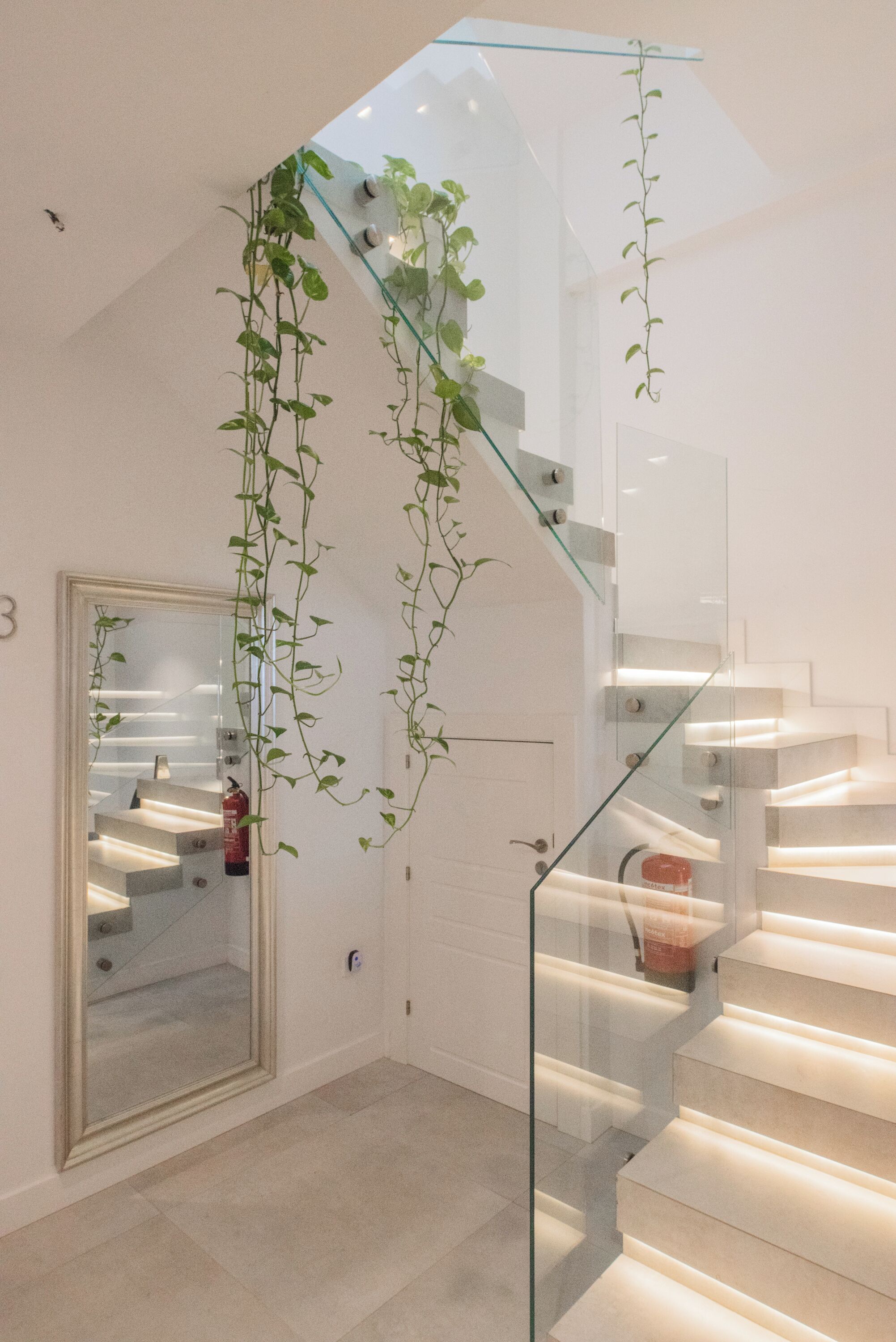 A modern interior with a glass staircase, a large mirror, and hanging plants.