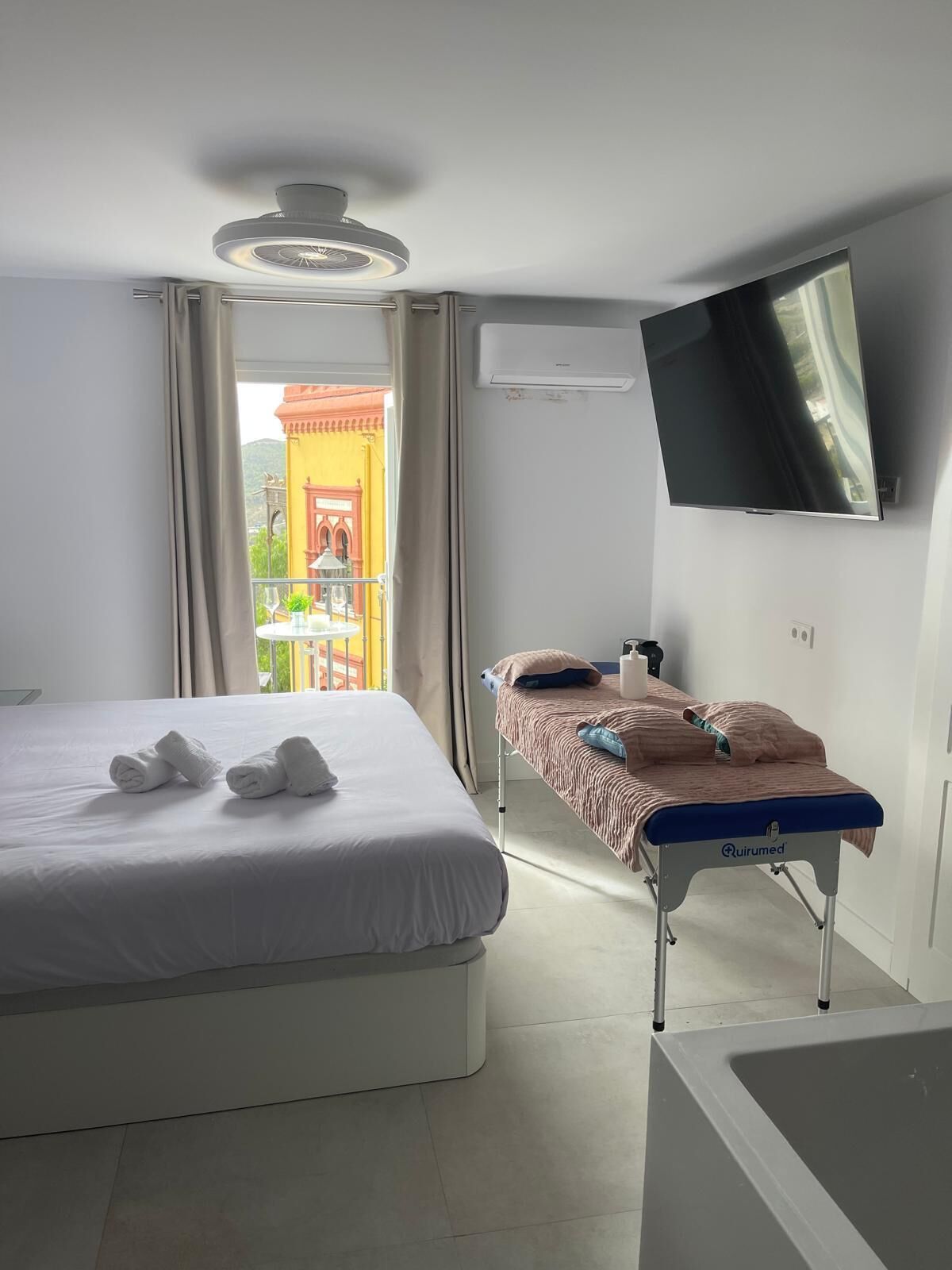 Una habitación de hotel con una cama, una mesa de masajes, un televisor y una ventana con cortinas.