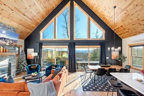 Amplia sala de estar con techo alto, ventanas grandes, chimenea y comedor con vistas al exterior.