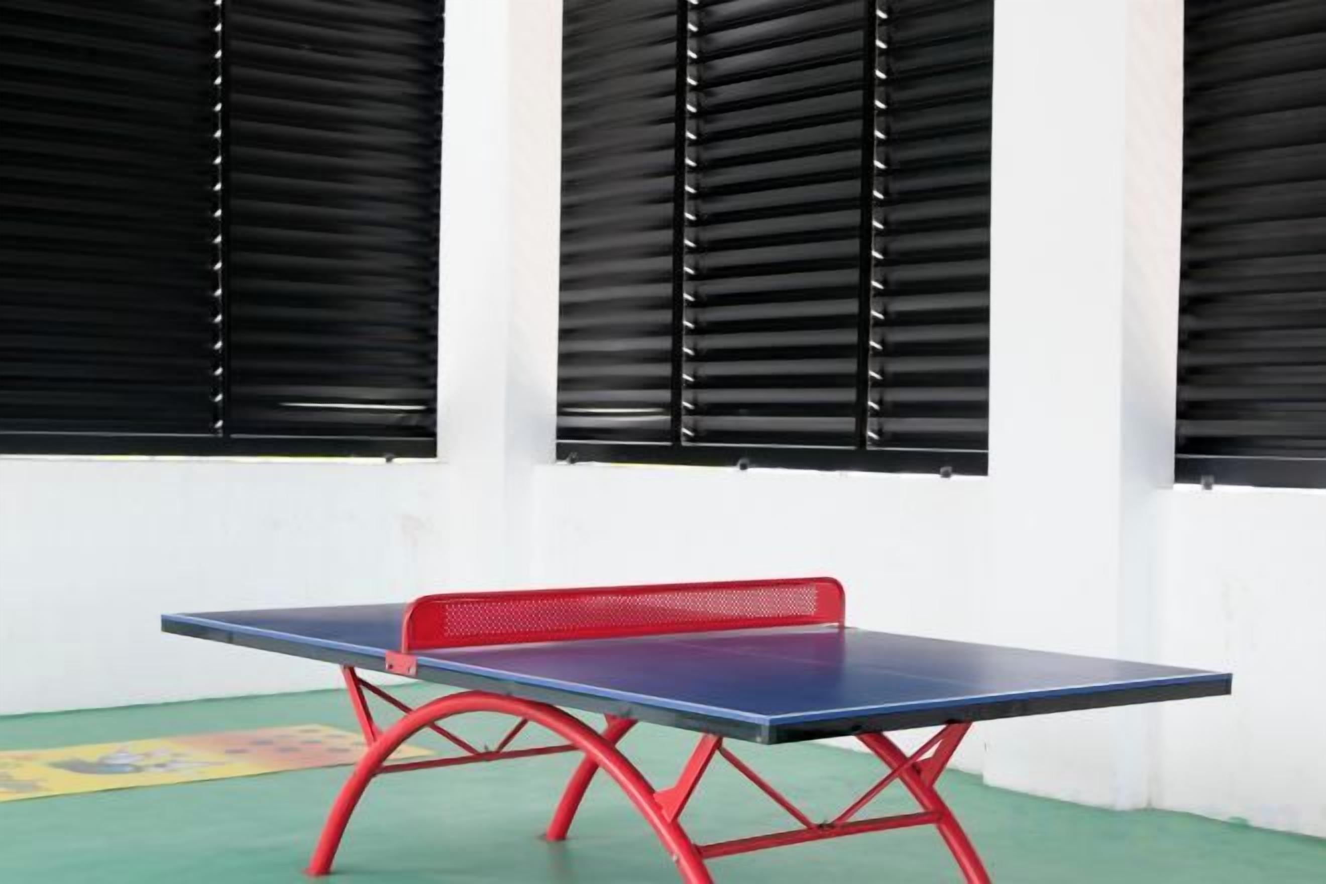 A ping pong table with a red frame, positioned in front of a wall with three black window blinds.
