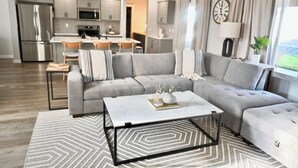 A modern living room with a grey sectional sofa, a coffee table, and a kitchen area in the background. - Ultimate Sports & Relaxation Retreat – Private Courts & Large Yard (Nampa)