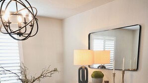 A dining area with a wooden table, chairs, a vase with branches, a lamp, a mirror, and a black cabinet with a plaid cloth. - Ultimate Sports & Relaxation Retreat – Private Courts & Large Yard (Nampa)