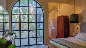 A bedroom with a large arched window, a bed, a wooden bench, a washing machine, and a hanging lamp. - Casa Toto - Centro Luxe Retreat  (Mérida)