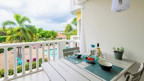 A balcony with a white railing, a table with a bottle, glasses, and a bowl of fruit, and a view of a swimming pool and palm trees.