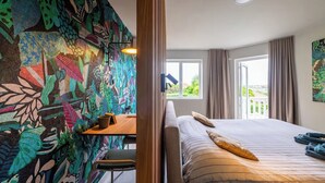 A bedroom with a bed, a desk, and a chair. There is a window with curtains and a view of greenery outside.