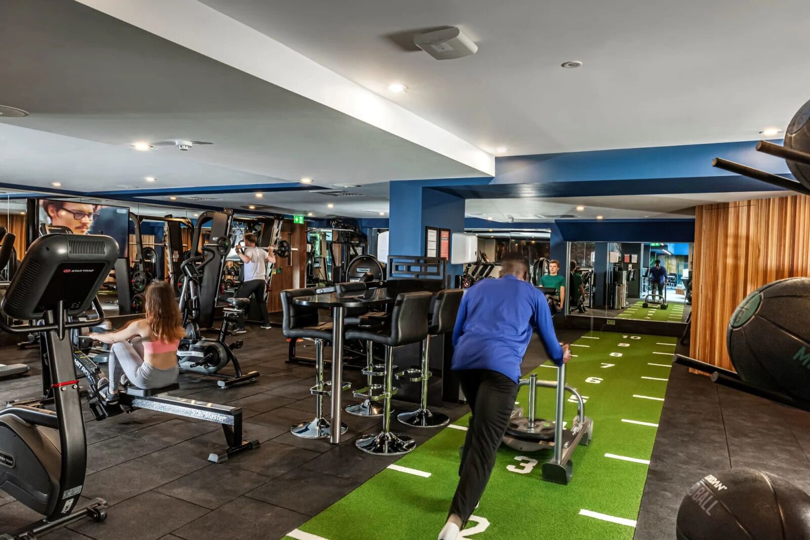 Une salle de sport équipée de divers appareils d’exercice, dotée d’une zone de tapis synthétique vert et d’un mur recouvert de miroirs.