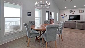 Ein geräumiges Wohnzimmer mit einem großen runden Tisch, grauen Polsterstühlen, einem Deckenventilator und einem Fernseher.