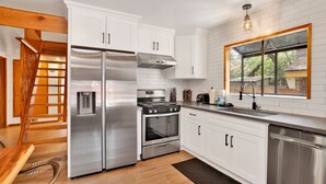A modern kitchen with white cabinetry, stainless steel appliances, a wooden staircase, and a window with a view of the outdoors. - Sugar Bear Pines #2493 (Sugarloaf)