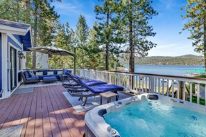 A deck with a hot tub, lounge chairs, and a sofa overlooking a lake. - Serenity Cove Lake House #2472 by Big Bear Vacations (Big Bear Lake)