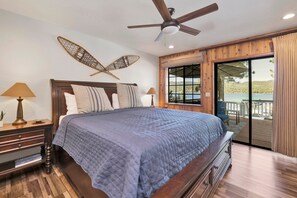 Un dormitorio con una cama grande, cabecera de madera, dos mesitas de noche con lámparas, una ventana con vista al mar y un balcón con mesa y sillas.