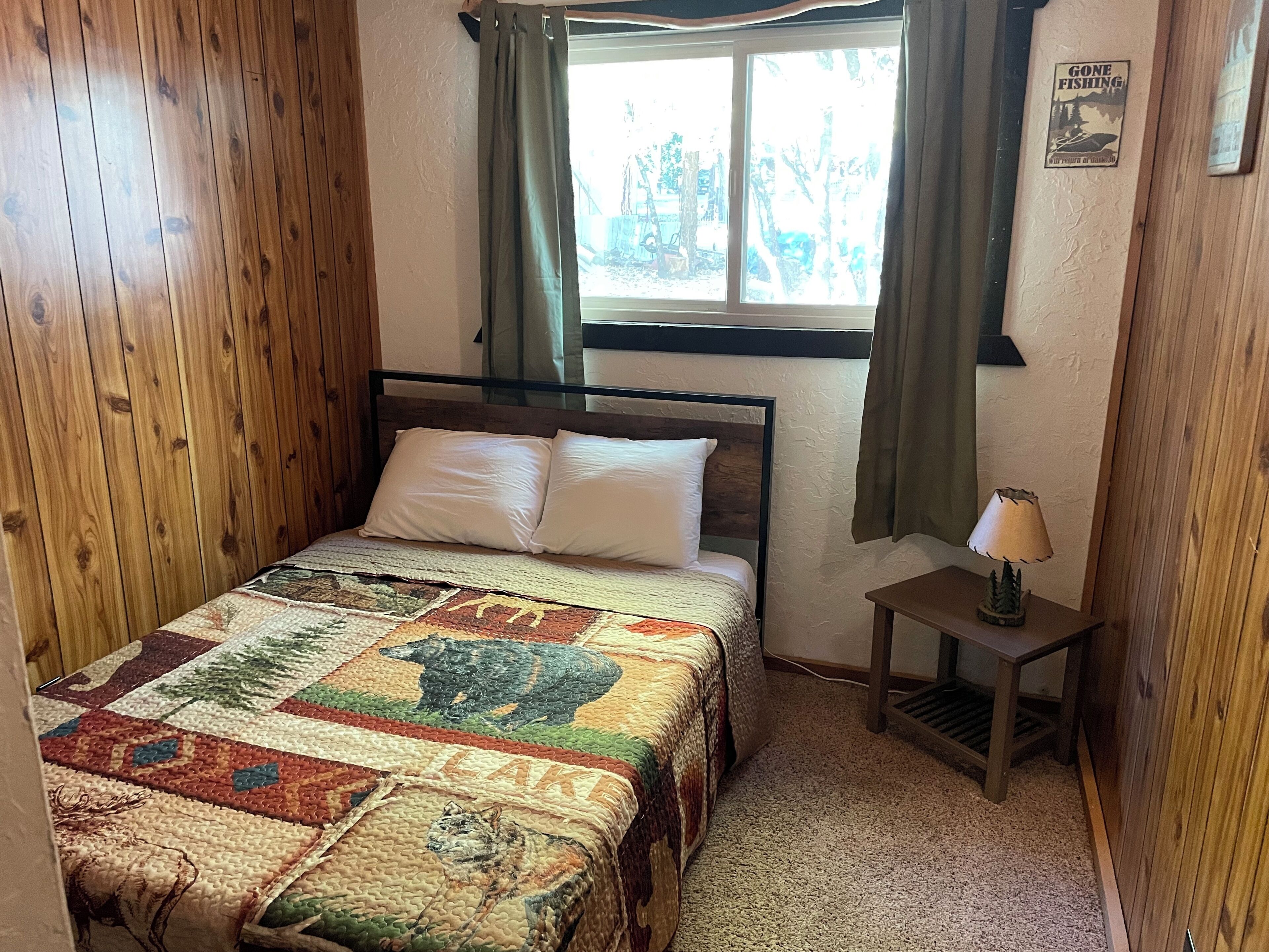 A bedroom with a wooden floor, a bed with a quilt, a nightstand with a lamp, and a window with curtains.