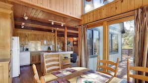A wooden cabin interior with a dining table, chairs, a kitchen area, and a large window.
