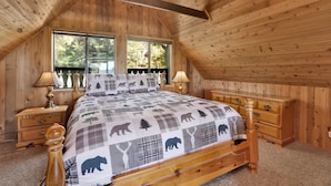 A bedroom with a sloped ceiling, wooden walls, a bed with a checkered blanket, two bedside tables, and a nightstand.