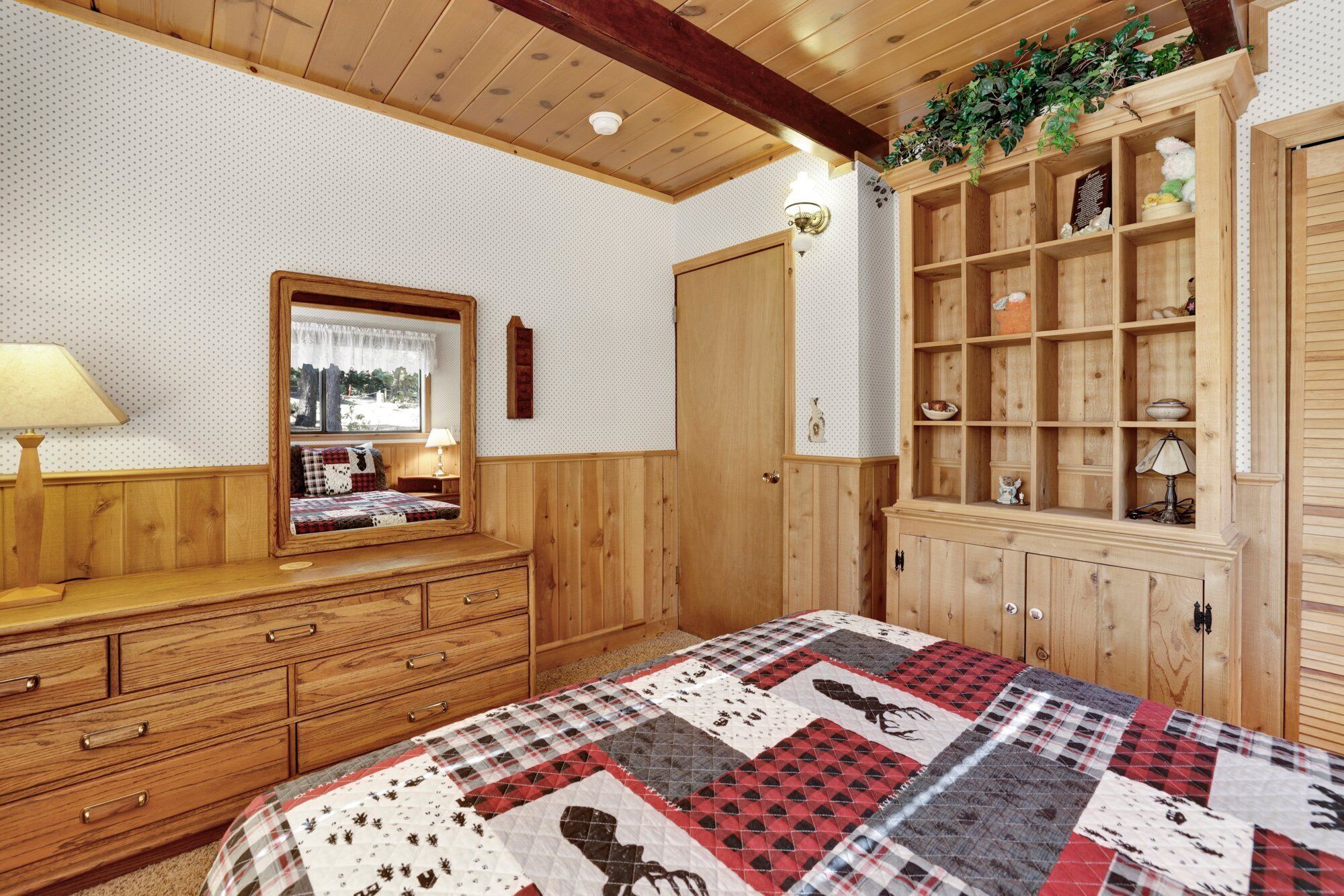 Un dormitorio acogedor con muebles de madera, una colcha acolchada, un aparador, un espejo y un armario de madera con estantes.