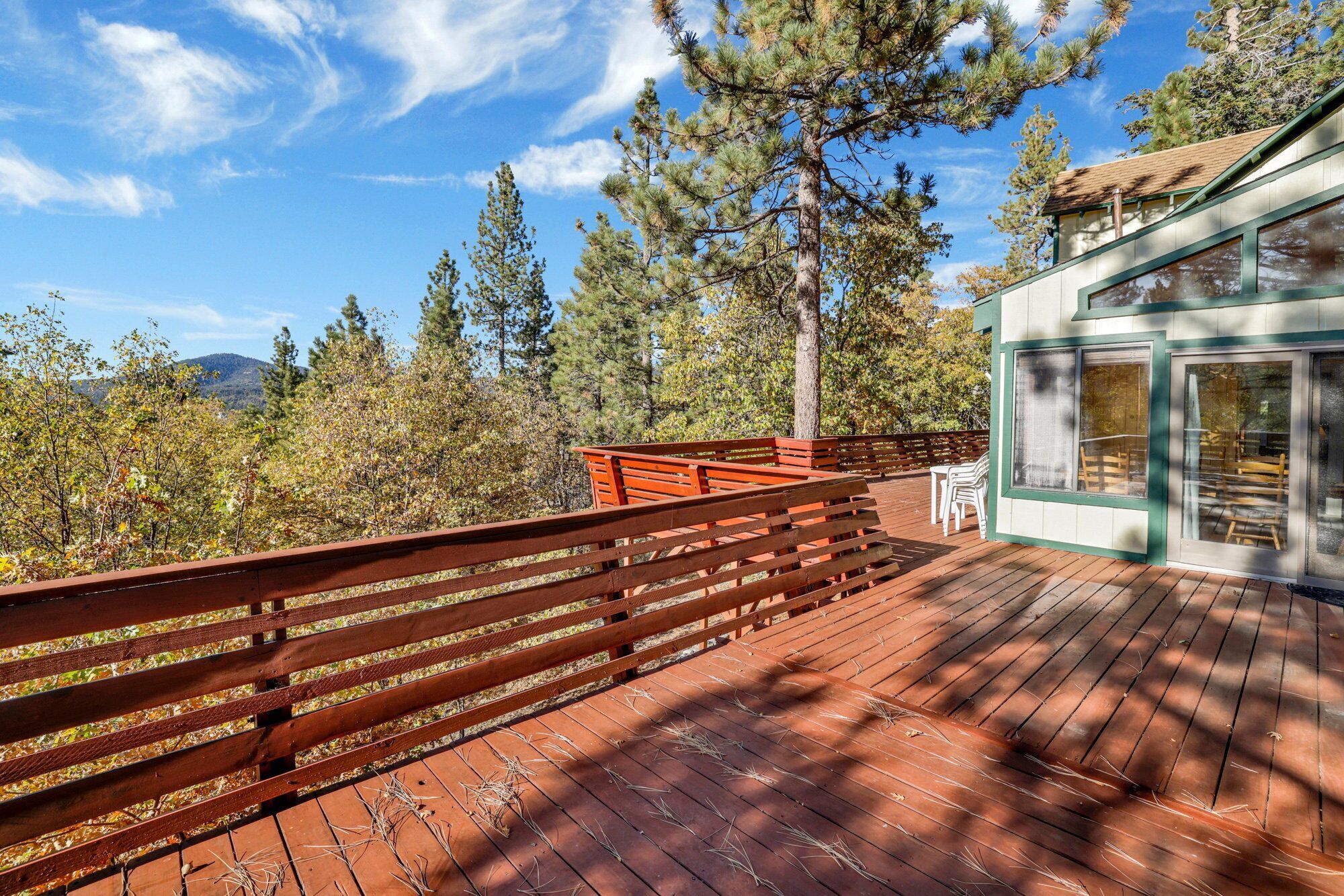 A wooden deck with a railing, overlooking a forested area with tall trees and a mountain in the distance.