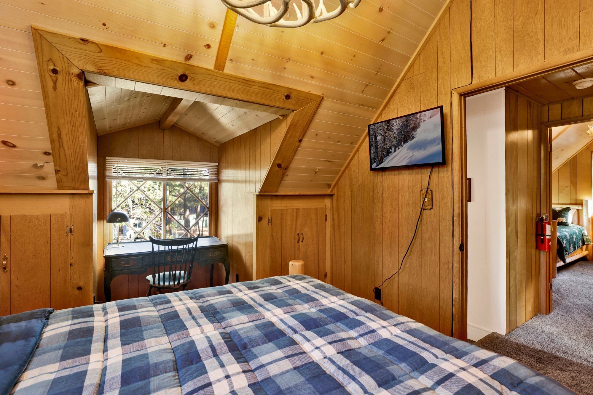 A cozy room with a wooden ceiling, a bed with a plaid blanket, a desk with a chair, and a window with blinds.