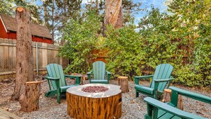 Un pozo de fuego circular de madera rodeado por cuatro sillas Adirondack verdes en un jardín con árboles y una cerca de madera al fondo.