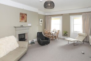 A living room with a fireplace, a sofa, a lounge chair, a dining table with chairs, and a large window with curtains. - Seaview Suite By My Getaways (Hinckley)