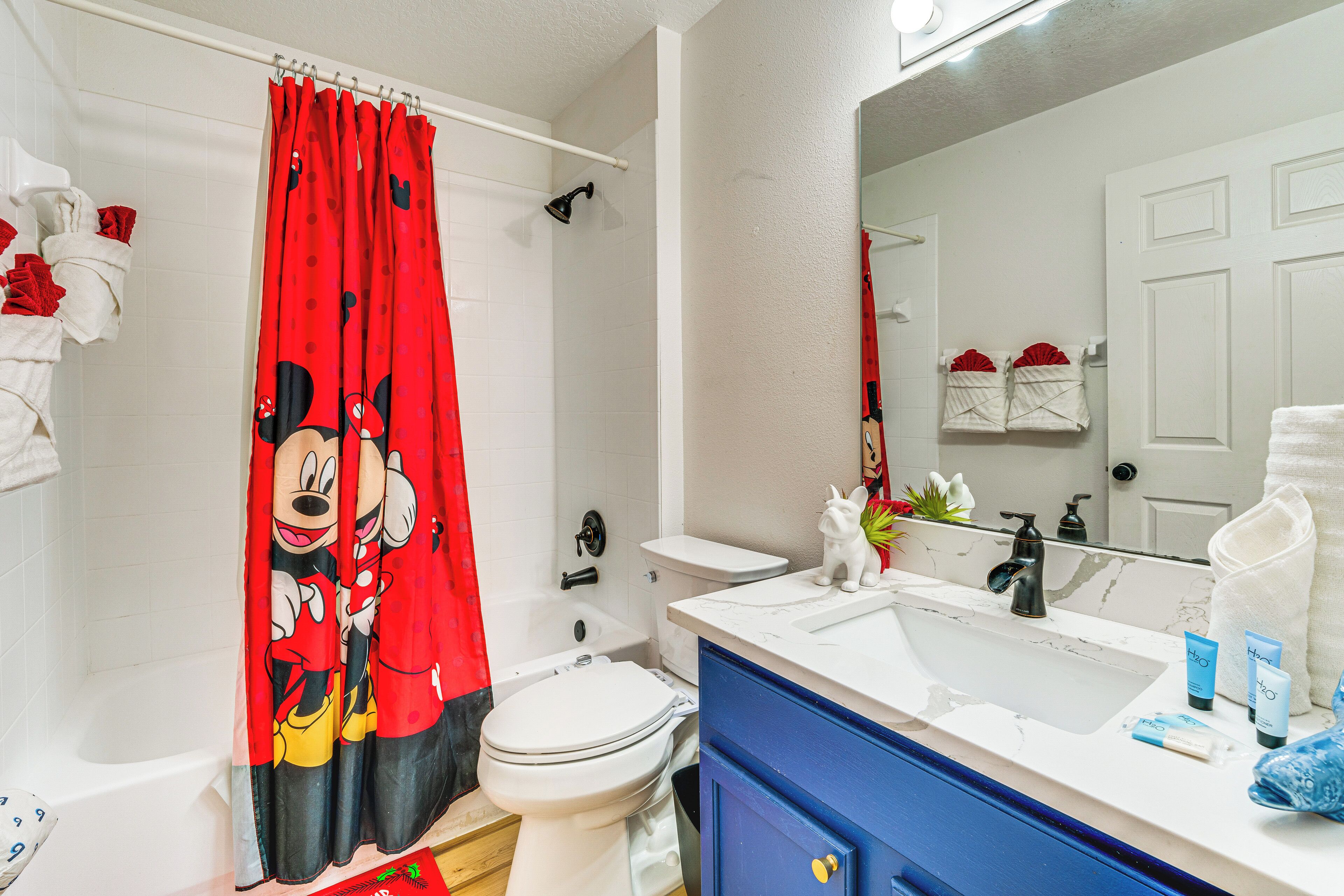 A bathroom with a Mickey Mouse shower curtain, a white toilet, a bathtub, a sink with toiletries, and a mirror reflecting the room.