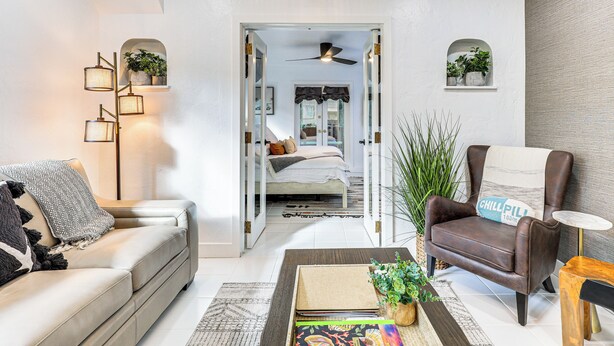 A modern living room with a sofa, armchair, and coffee table, featuring a view into a bedroom with a bed and a ceiling fan. - 'The Peacock Bungalow' Near Fort Lauderdale Beach! (Fort Lauderdale)