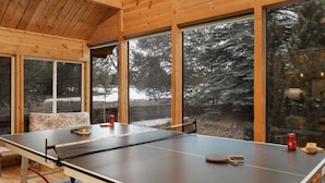 A ping pong table in a room with large windows, wooden walls, and a view of trees and snow outside. - Come stay at the Moose Jam! (Island Park)