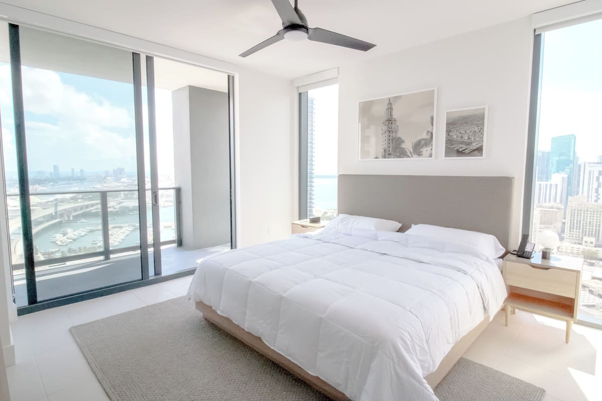 A modern bedroom with a large bed, a nightstand, and a view of a cityscape through floor-to-ceiling windows.