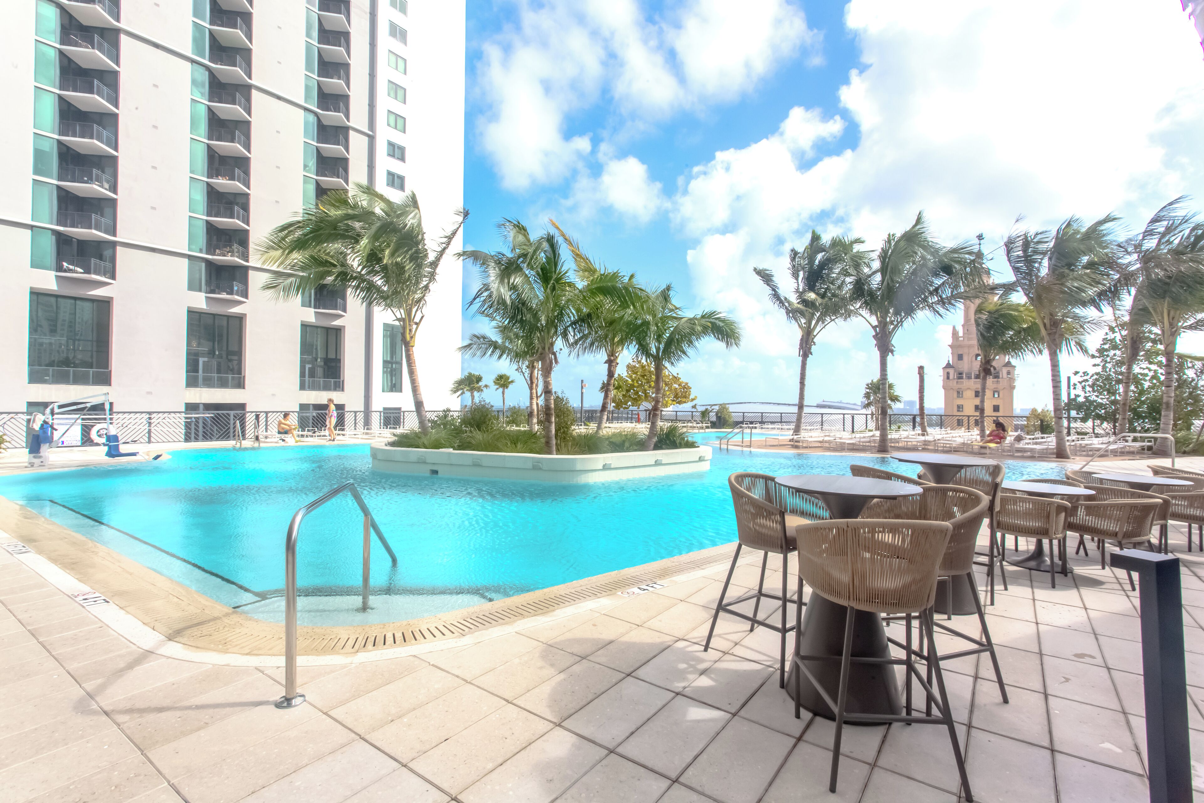 A swimming pool with a poolside area, palm trees, and a tall building in the background.