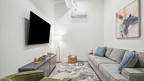 A modern living room with a flat-screen TV, a grey sofa with patterned cushions, a green armchair, a woven basket, and a floor lamp. - Centrally located, unique and brand new condo (Houston)