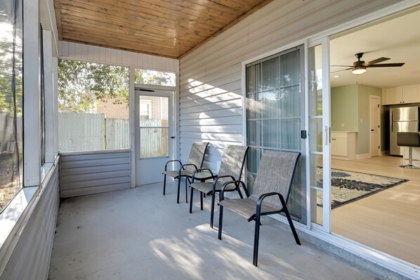 A covered porch with four chairs, a sliding glass door leading to a kitchen, and a ceiling fan. - 1 Mi to Pier: Screened Patio at Murrells Inlet Apt (Murrells Inlet)