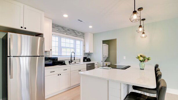 A modern kitchen with white cabinets, a stainless steel refrigerator, a dining area with a white island and black chairs, and pendant lighting. - 1 Mi to Pier: Screened Patio at Murrells Inlet Apt (Murrells Inlet)