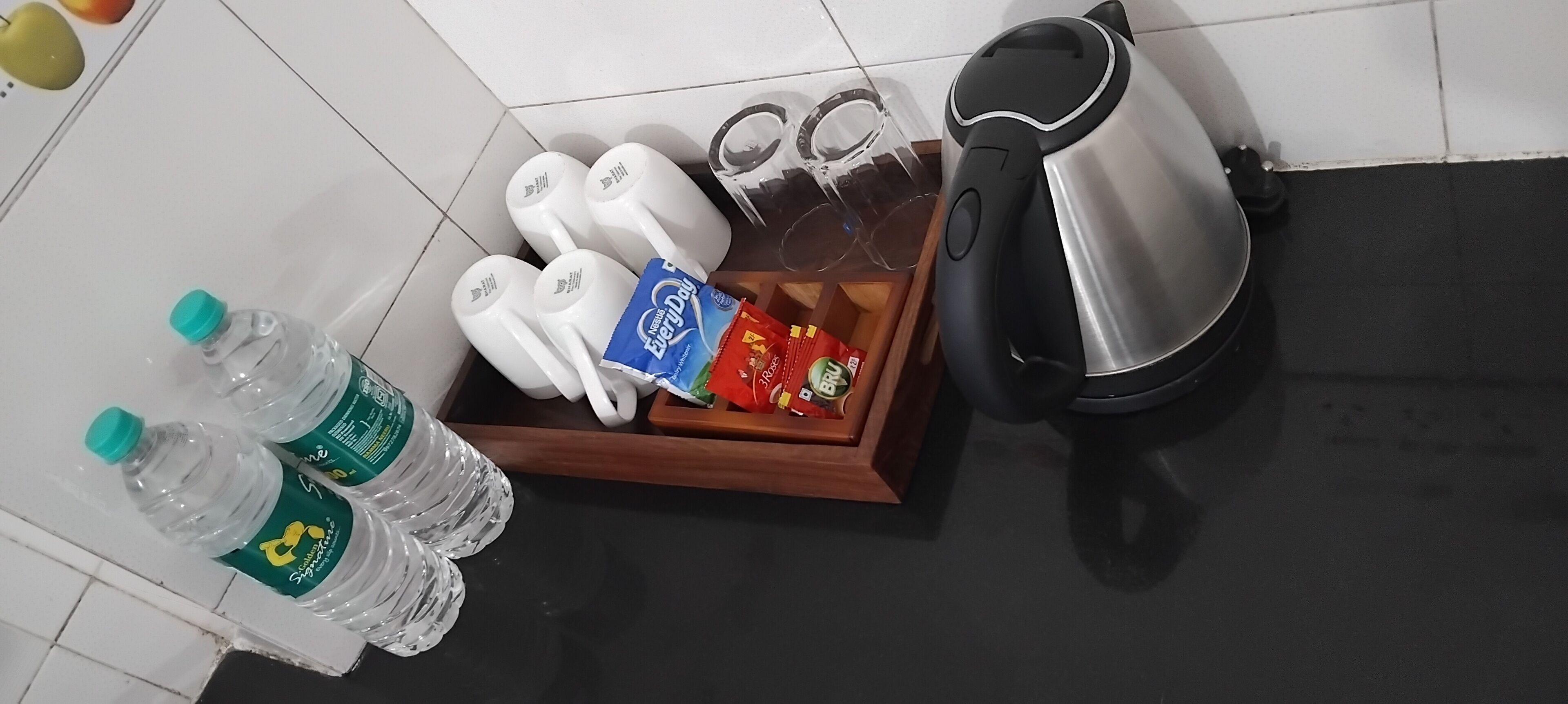 Un comptoir de cuisine avec une bouilloire, quatre tasses blanches, une boîte de sachets de thé et plusieurs bouteilles d’eau.