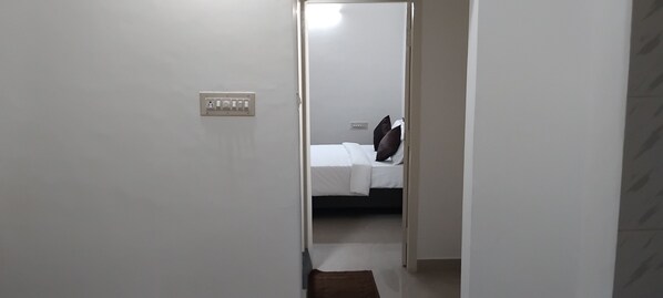 A narrow hallway leading to a bedroom with a bed, a nightstand, and a wall socket. - SLV Aashraya Apartment Stay (Mysore)