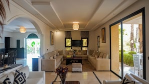 Amplia sala de estar con un conjunto de sofá, una mesa de centro y un televisor montado en la pared. Se aprecia una ventana grande con vista a palmeras y un balcón.