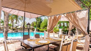 A poolside dining area with a table set for two, surrounded by lounge chairs, under a shaded canopy.