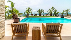 A poolside area with wooden chairs and tables, surrounded by potted plants and umbrellas, overlooking a clear blue pool and distant landscape.