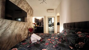 Habitación de hotel con una cama grande, un televisor de pantalla plana montado en una pared con textura, una pequeña mesa con una silla y vistas al área de la cocina.