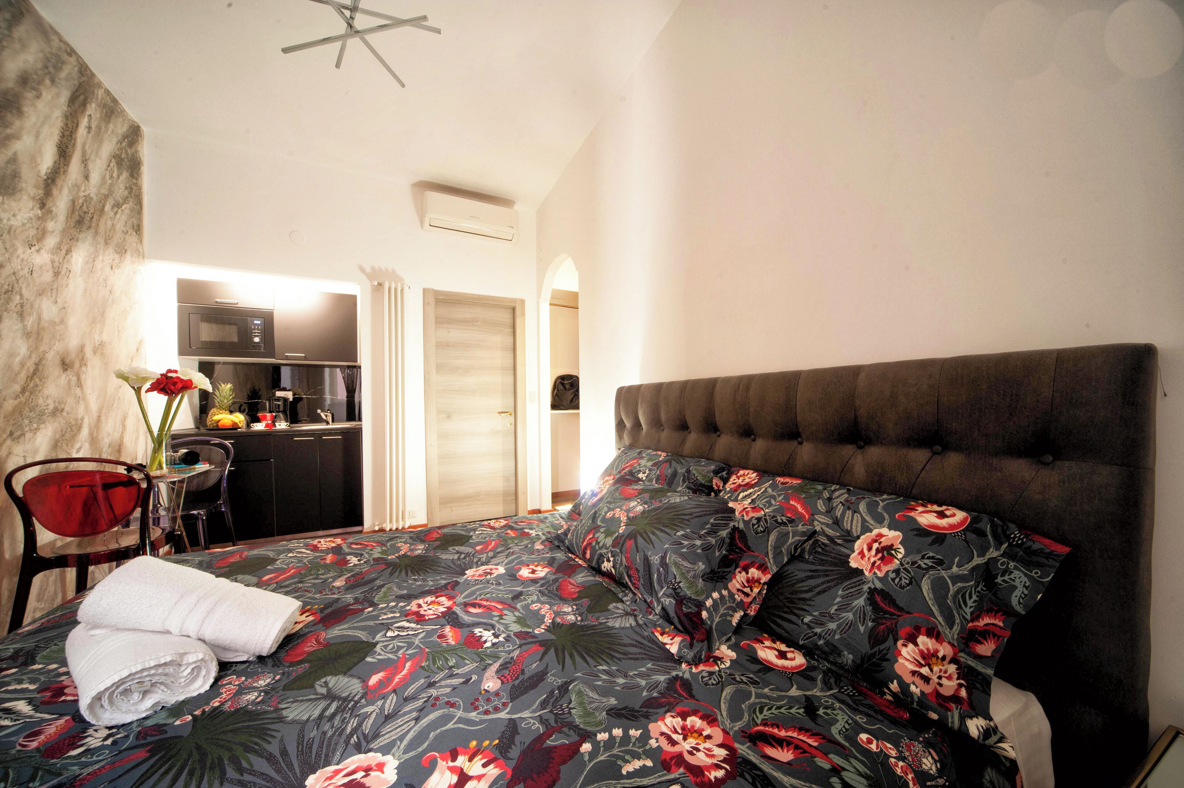 Quarto com cama, uma cadeira vermelha, uma mesinha e uma kitchenette ao fundo.