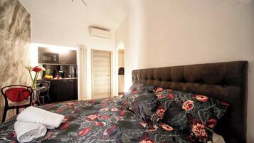 Quarto com cama, uma cadeira vermelha, uma mesinha e uma kitchenette ao fundo.