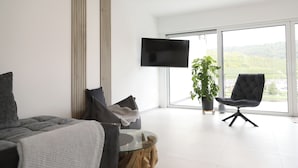 A modern living room with a sofa, a glass coffee table, a black chair, a flat-screen TV, and a large window with a view of greenery.