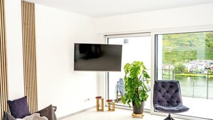 A modern living room with a flat-screen TV, a large potted plant, a tufted chair, and a sofa with a throw blanket.