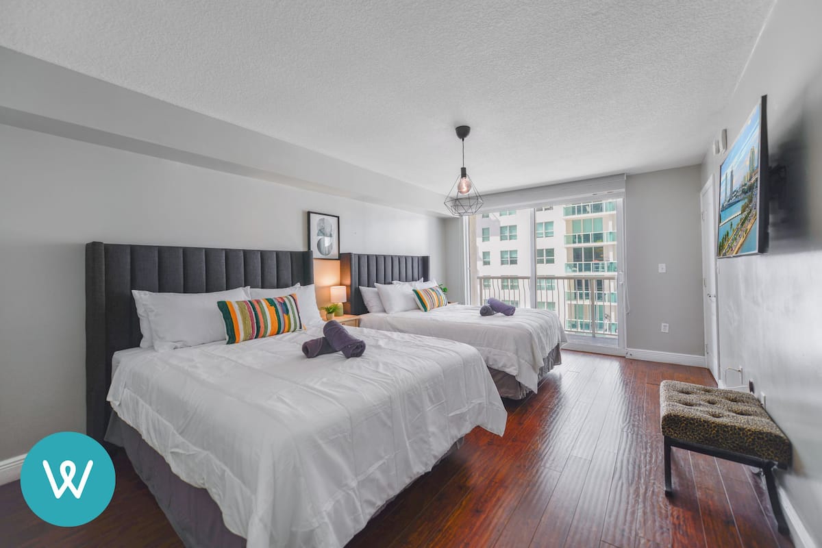 Habitación de hotel con dos camas, un banco y un amplio ventanal con vistas a la ciudad.