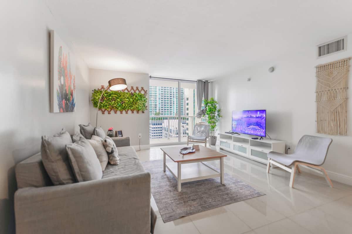 Una sala de estar moderna con sofá, mesa de centro, televisor y un amplio ventanal con vista a la ciudad.
