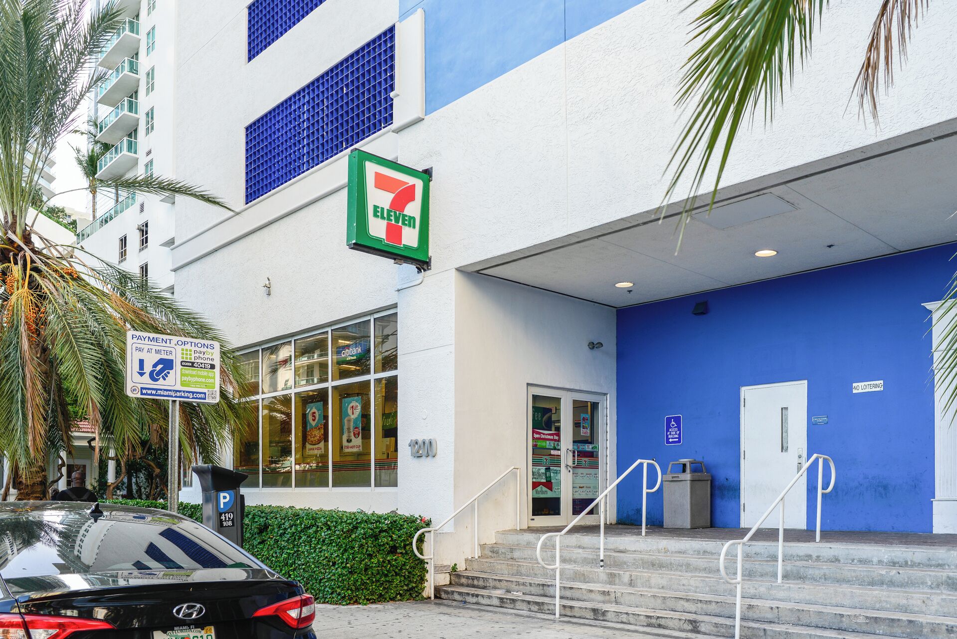 Un magasin 7-Eleven, avec une façade bleue, un panneau indiquant un espace de stationnement et un escalier menant à l’entrée.