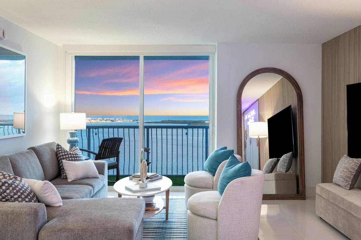 Una habitación de hotel moderna con balcón, un ventanal grande con vistas a la ciudad, una mesa de centro redonda y una zona de estar con sofás y sillones.