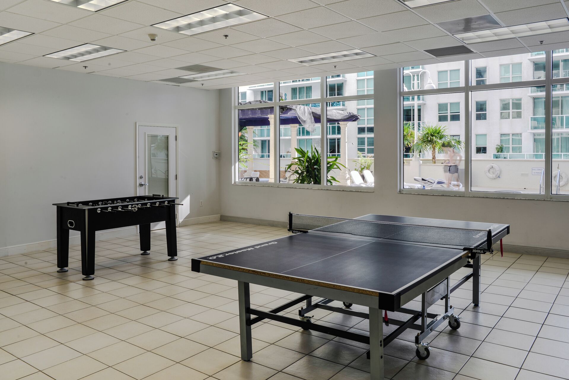 Una stanza con tavolo da ping pong, tavolo da calcio e ampie finestre vista su un edificio e piante da esterno.