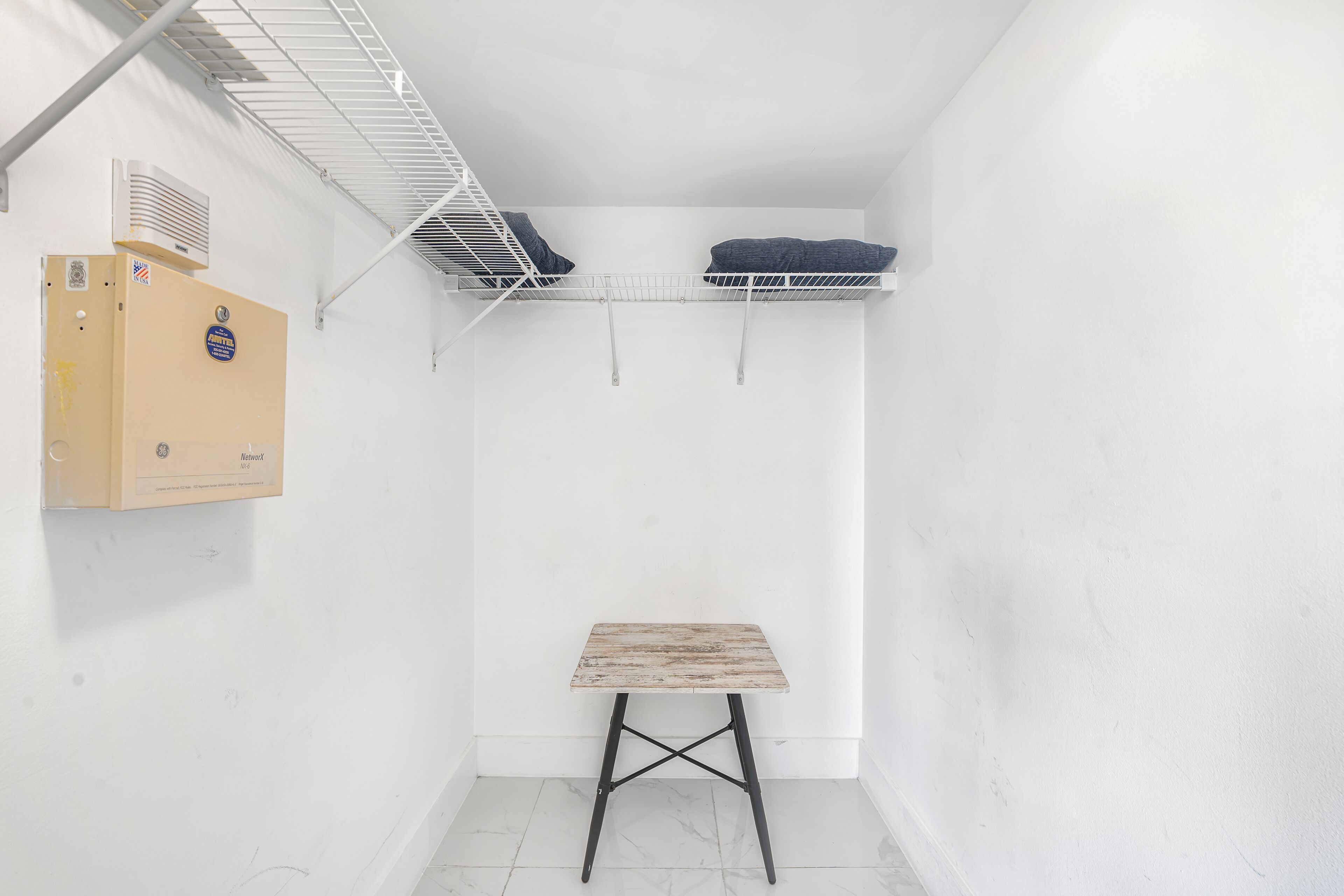 Une petite pièce aux murs blancs, avec une table en bois, une étagère blanche sur laquelle reposent deux coussins et une boîte fixée au mur.