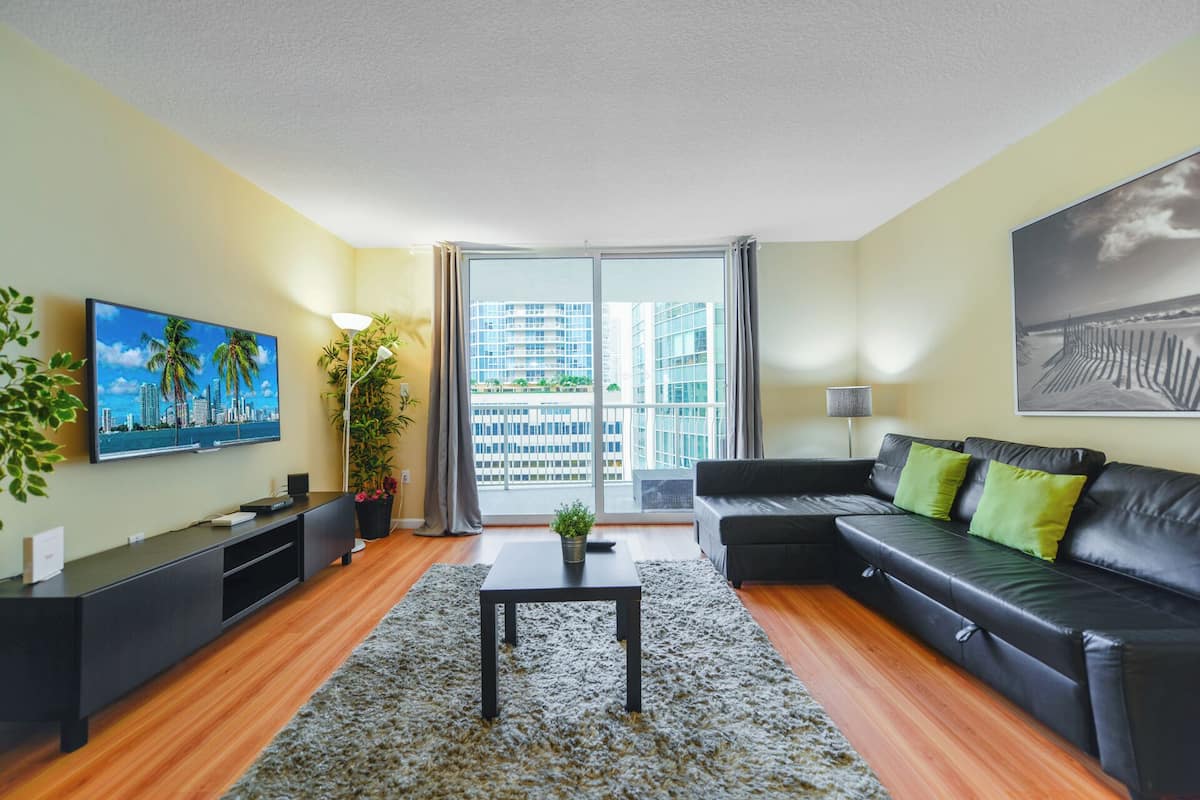 Una sala de estar moderna con un sofá de cuero negro, una mesita de centro, un televisor en la pared, una planta en maceta y vistas a un paisaje urbano a través de un ventanal grande.