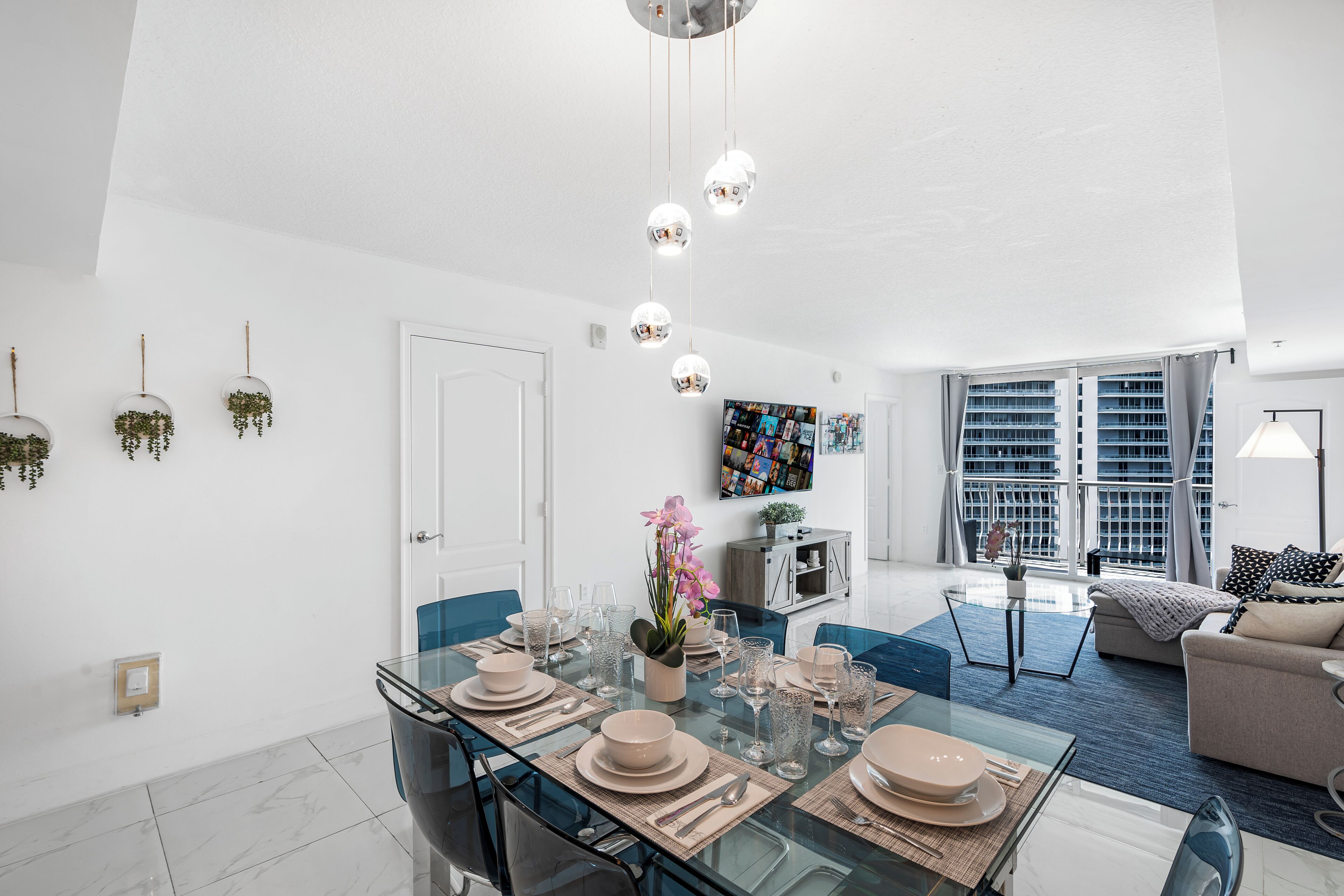 A modern living room with a glass dining table set for six, a sofa, and a TV mounted on the wall.
