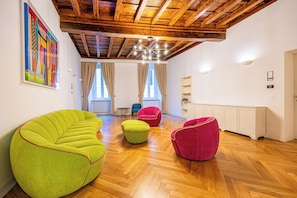 Une pièce spacieuse avec un plancher en bois, un plafond haut avec des poutres apparentes et une grande fenêtre avec des rideaux. On y trouve plusieurs chaises colorées et un tableau accroché au mur.